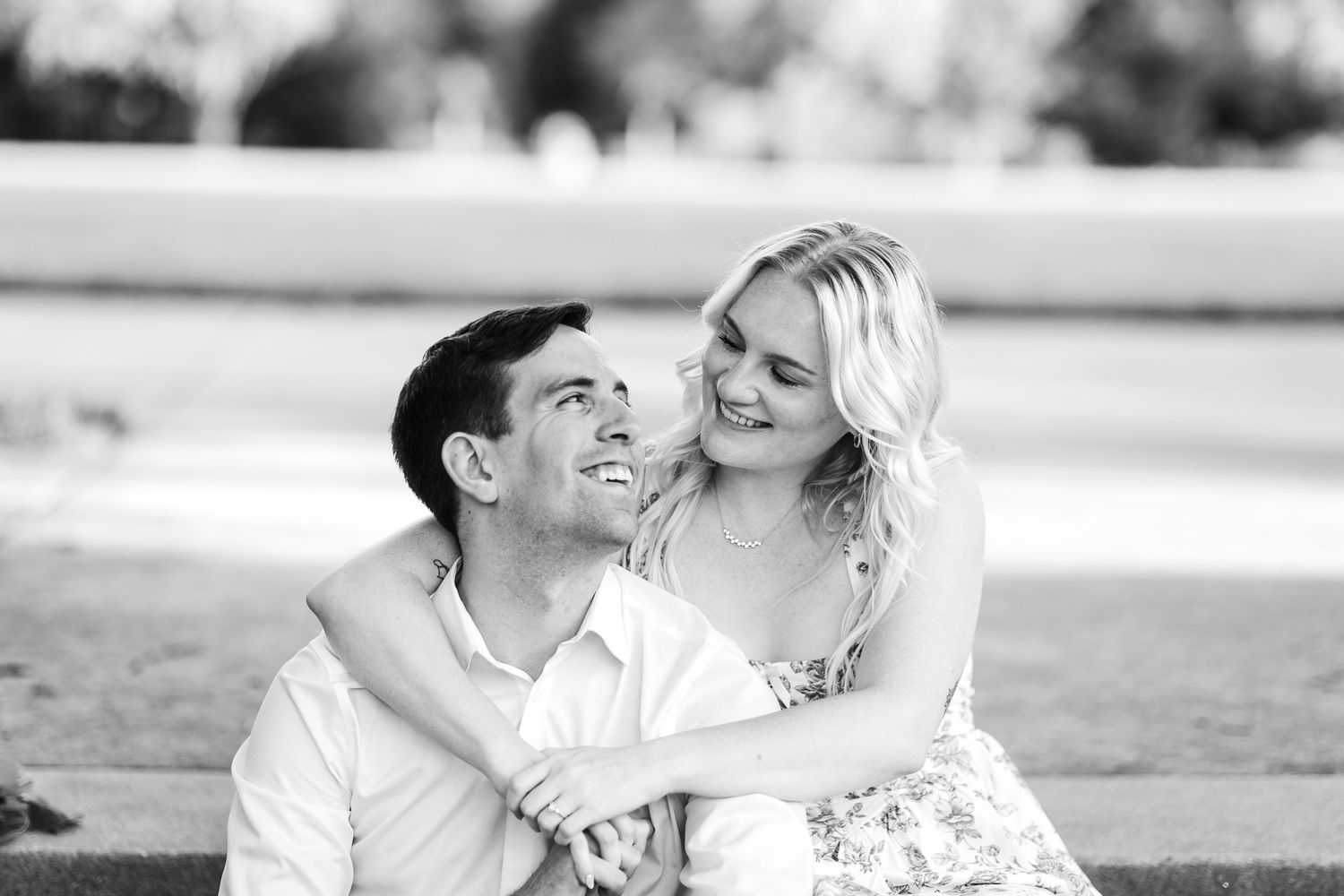 Happy couple sitting outdoors in black and white photo, woman embracing man from behind as they smile.