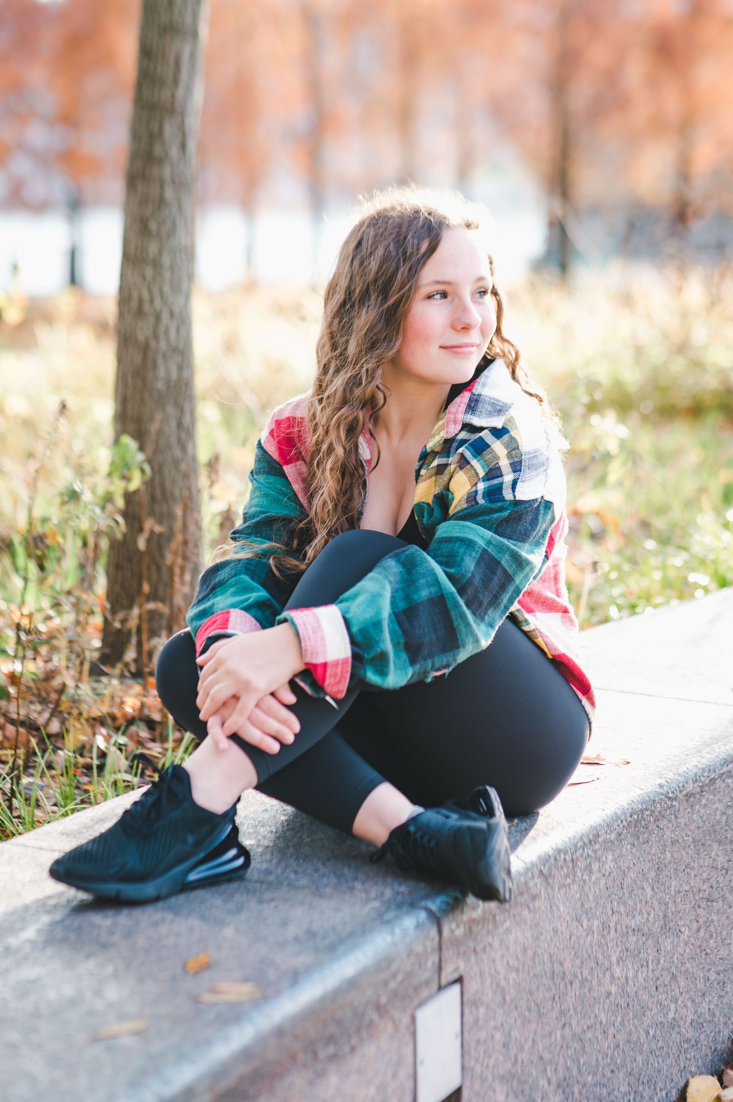 Teen girl in colorful plaid jacket sitting on stone ledge in autumn park setting.