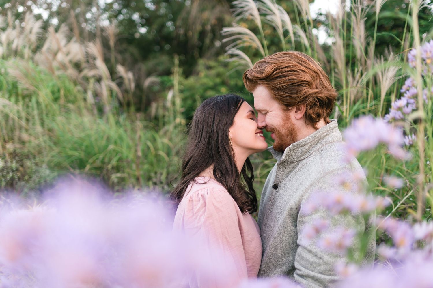 Romantic couple nearly kissing surrounded by purple wildflowers and tall ornamental grasses in garden.