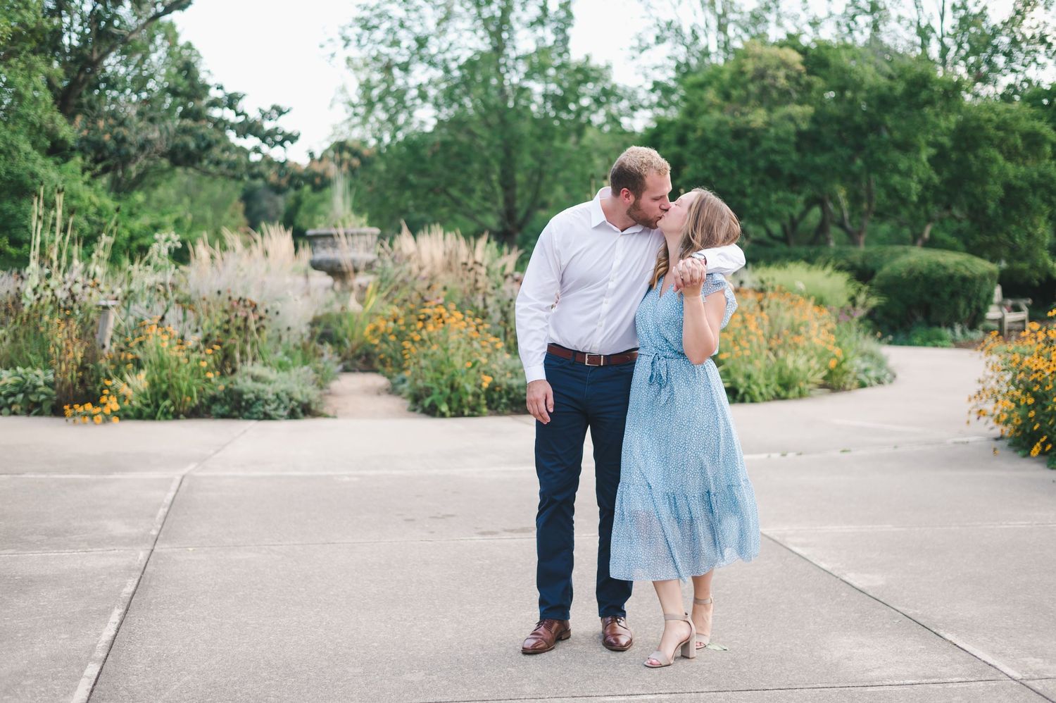 Couple shares a kiss on garden pathway surrounded by colorful flowers, woman in light blue dress.