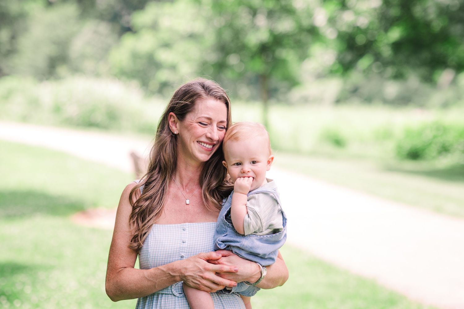 A smiling mother in a grey dress holds her baby boy in a sunny park pathway.