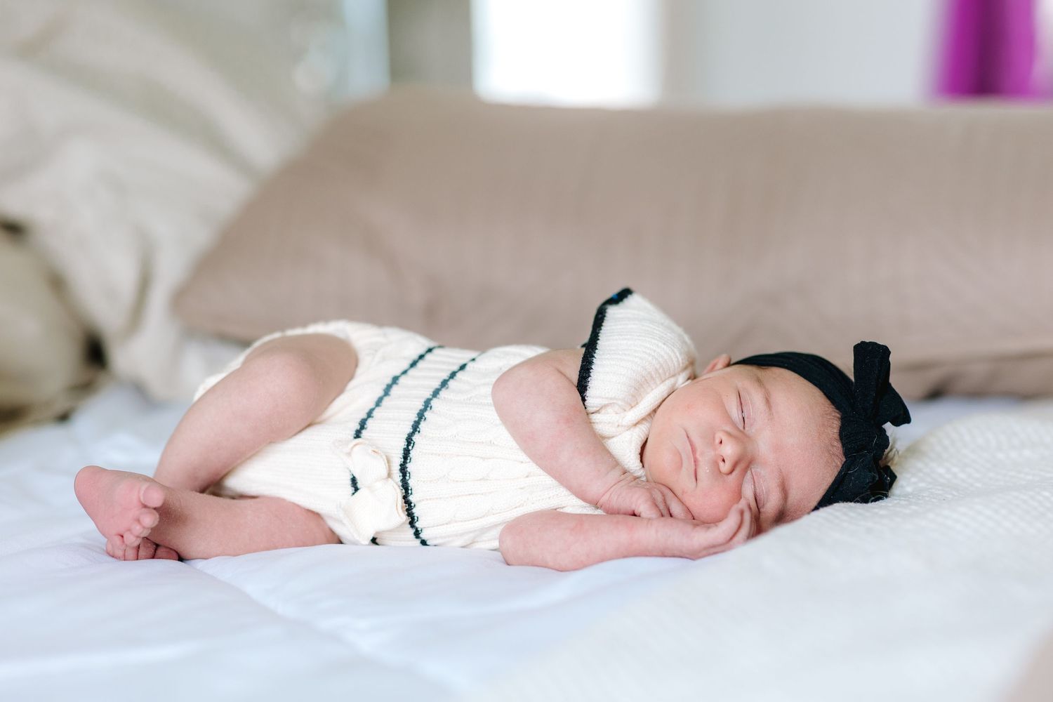 Peaceful newborn baby in white knit outfit sleeping with black bow headband on white bedding.
