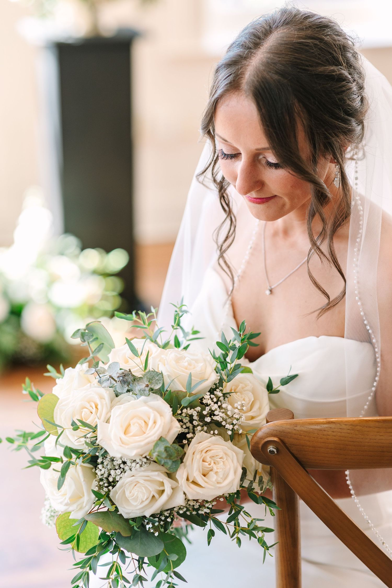 Elegant bride in white gown and veil gazing at her white rose bridal bouquet.