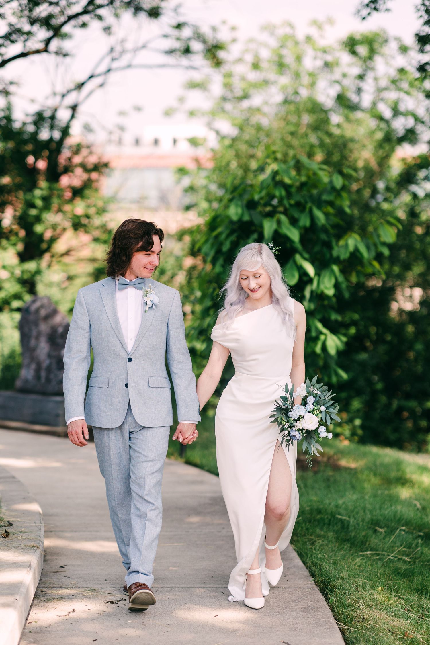 Couple walks hand in hand on sunlit garden path, bride in sleek white dress carrying white floral bouquet.