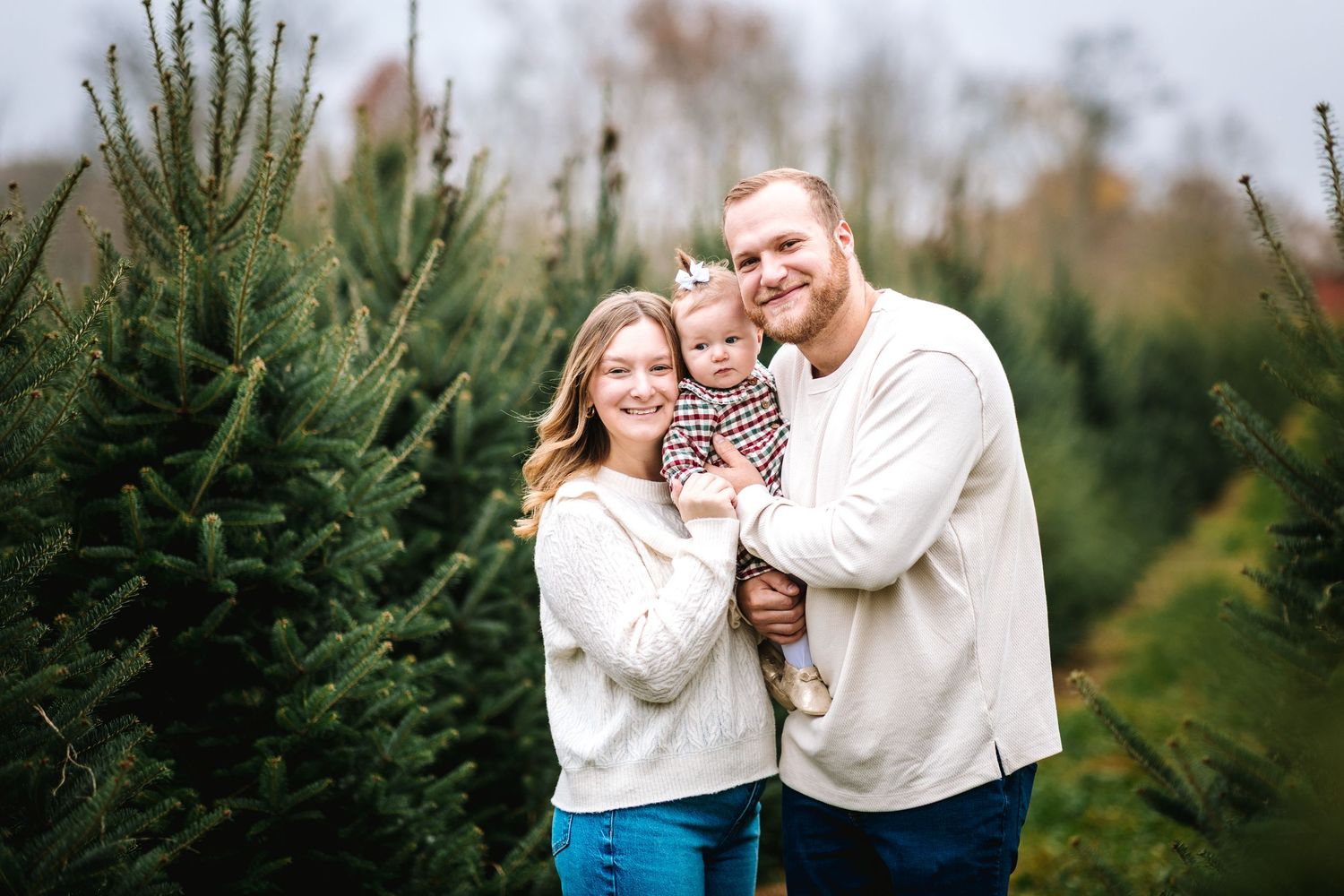 Young family with baby poses among green Christmas trees at a tree farm.