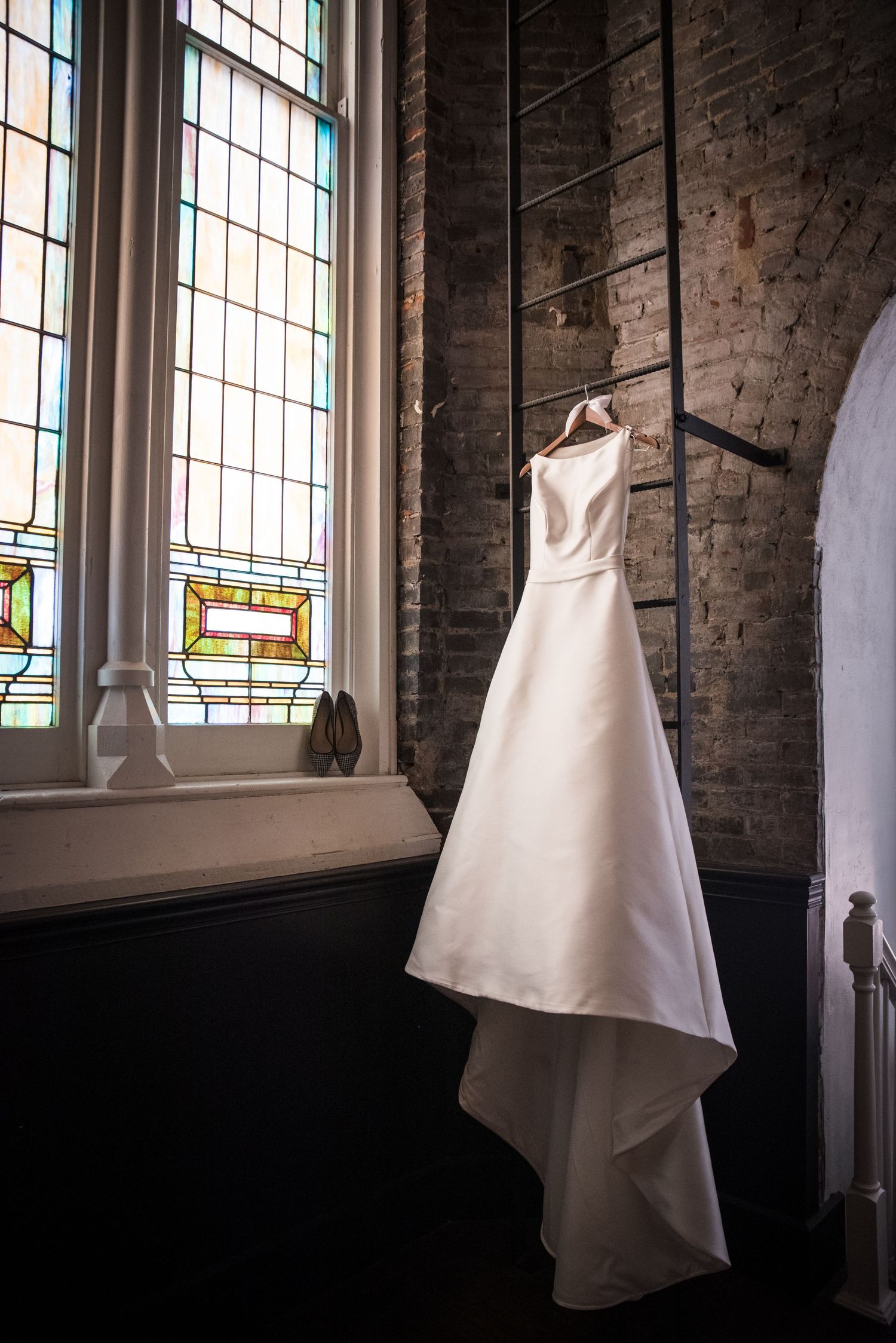 White satin A-line wedding gown hanging on hanger near stained glass window in rustic brick church interior at the transept.