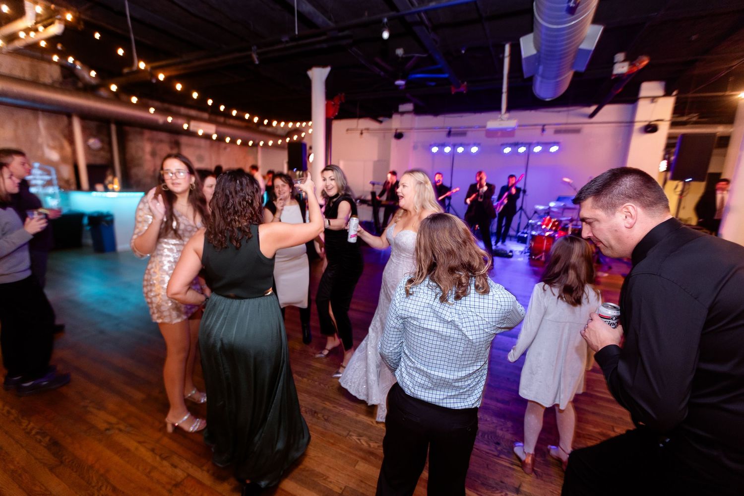 Wedding reception guests dancing together on a hardwood floor with a live band performing on a lit stage.