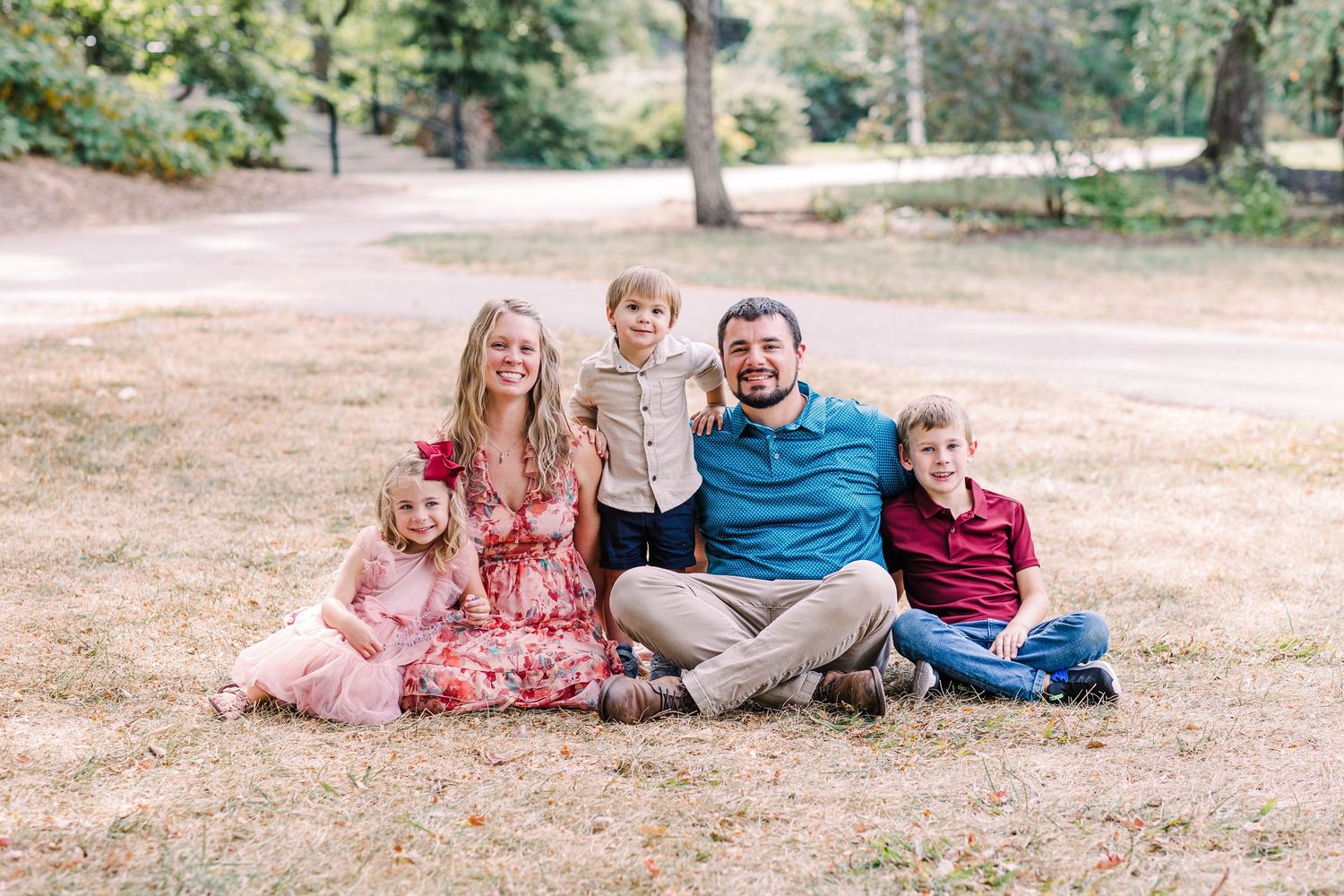 Family of five sitting on grass in a park, parents with three young children smiling for a portrait.
