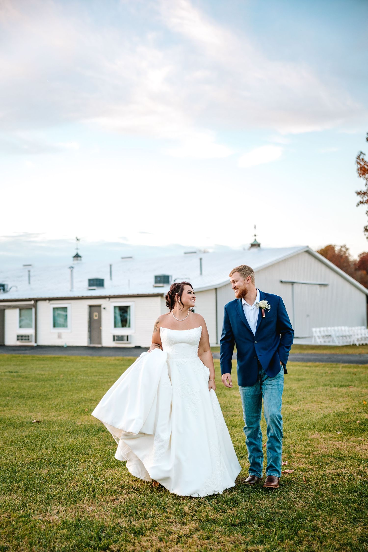 Bride in a flowing white gown and groom in a blue suit walk together on a green farm lawn at sunset.