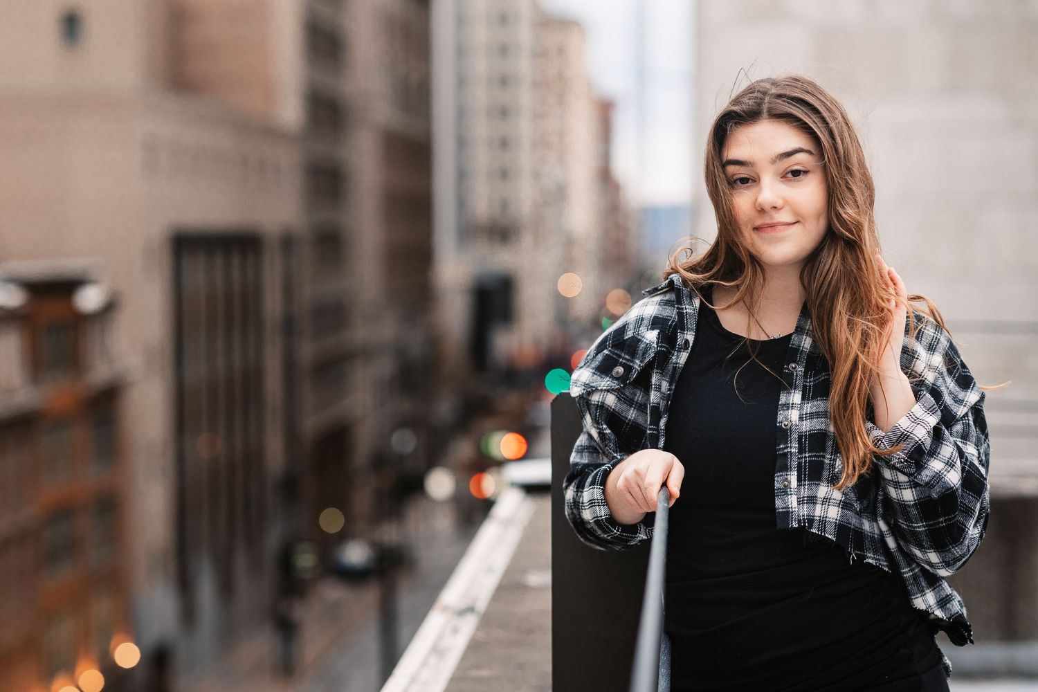 Young woman in plaid shirt leaning on a railing with a blurred city skyline behind her.