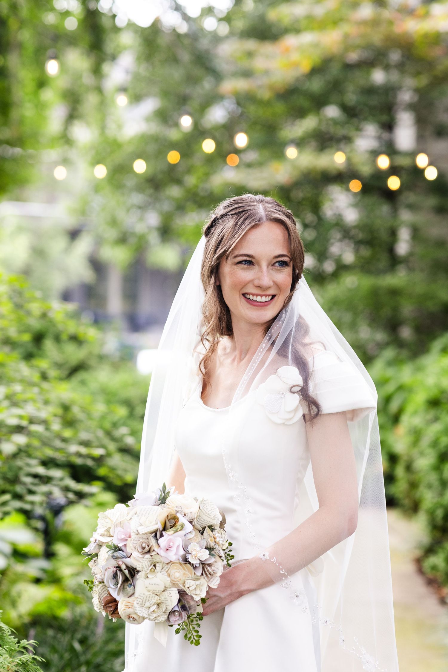 Smiling bride in white gown and veil holding bouquet in lush garden setting.