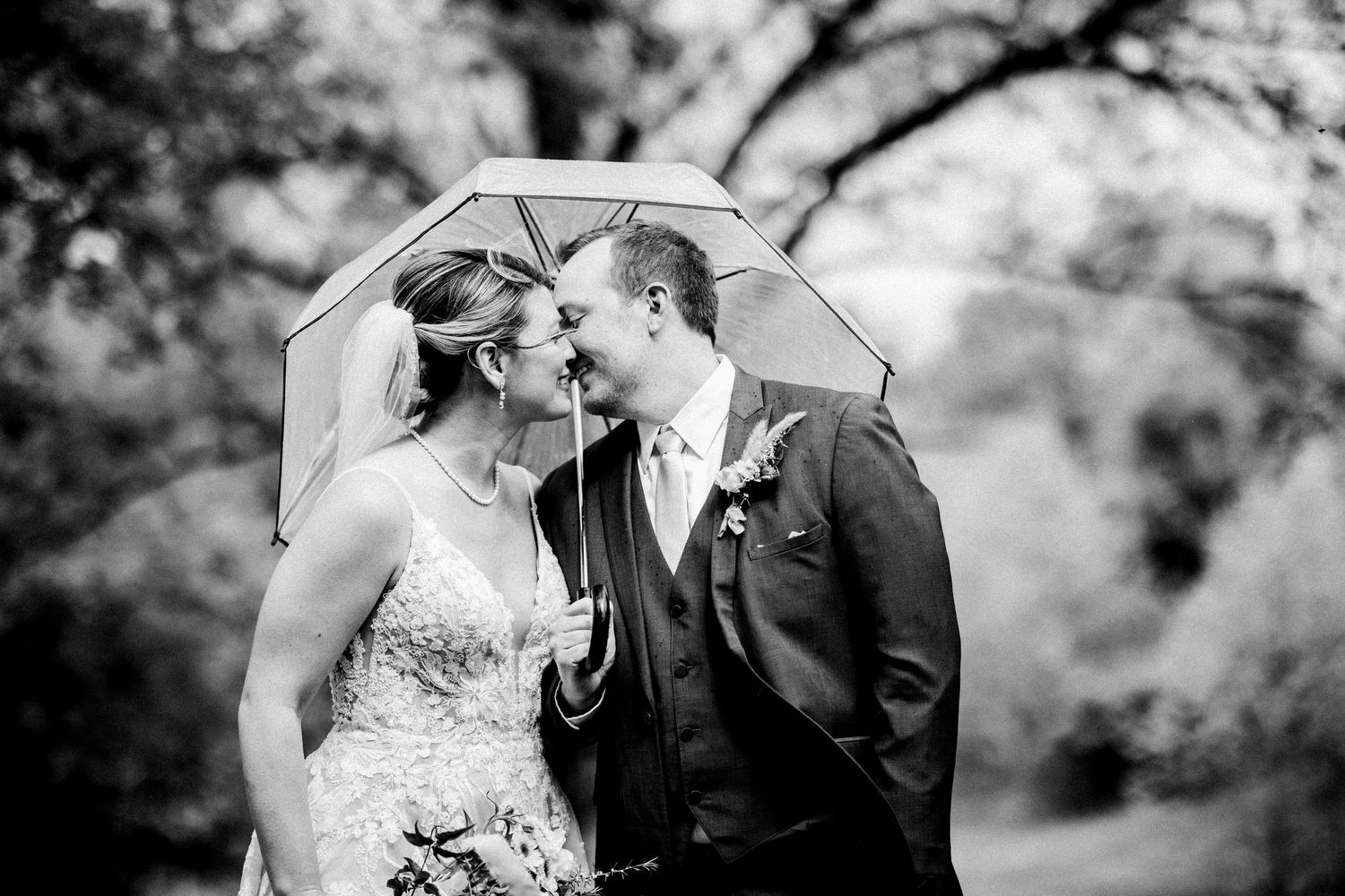 Black and white photo of bride and groom sharing a kiss under a clear umbrella in the rain.