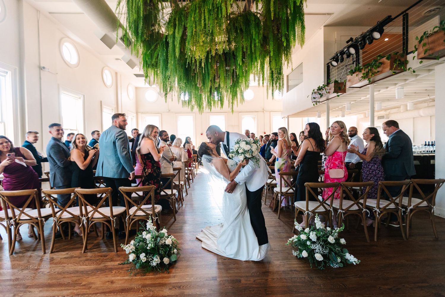 Bride and groom sharing their first kiss during a wedding ceremony beneath hanging greenery at the alcove by madtree.