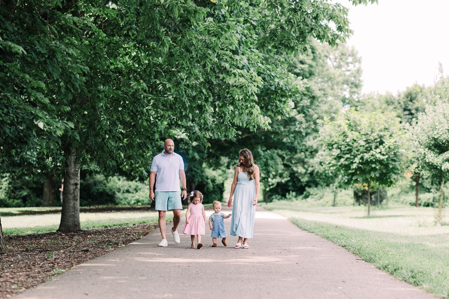A family of four walks hand-in-hand down a tree-lined park path on a sunny day.