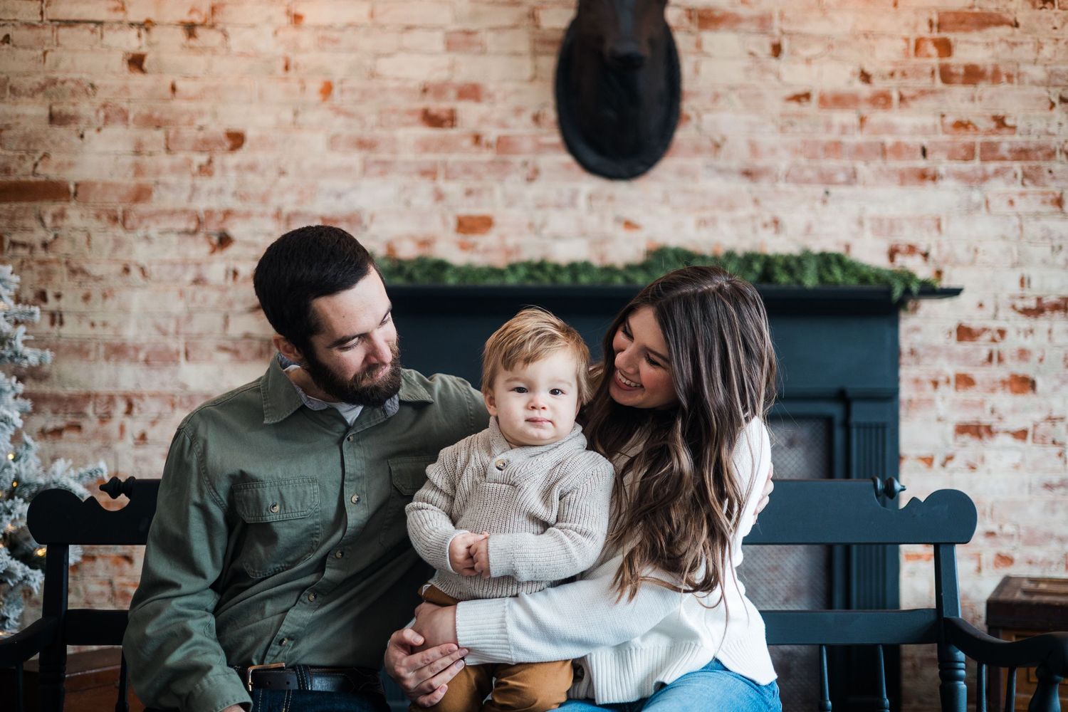 A young family with a toddler sits together on a bench in front of a rustic brick fireplace.