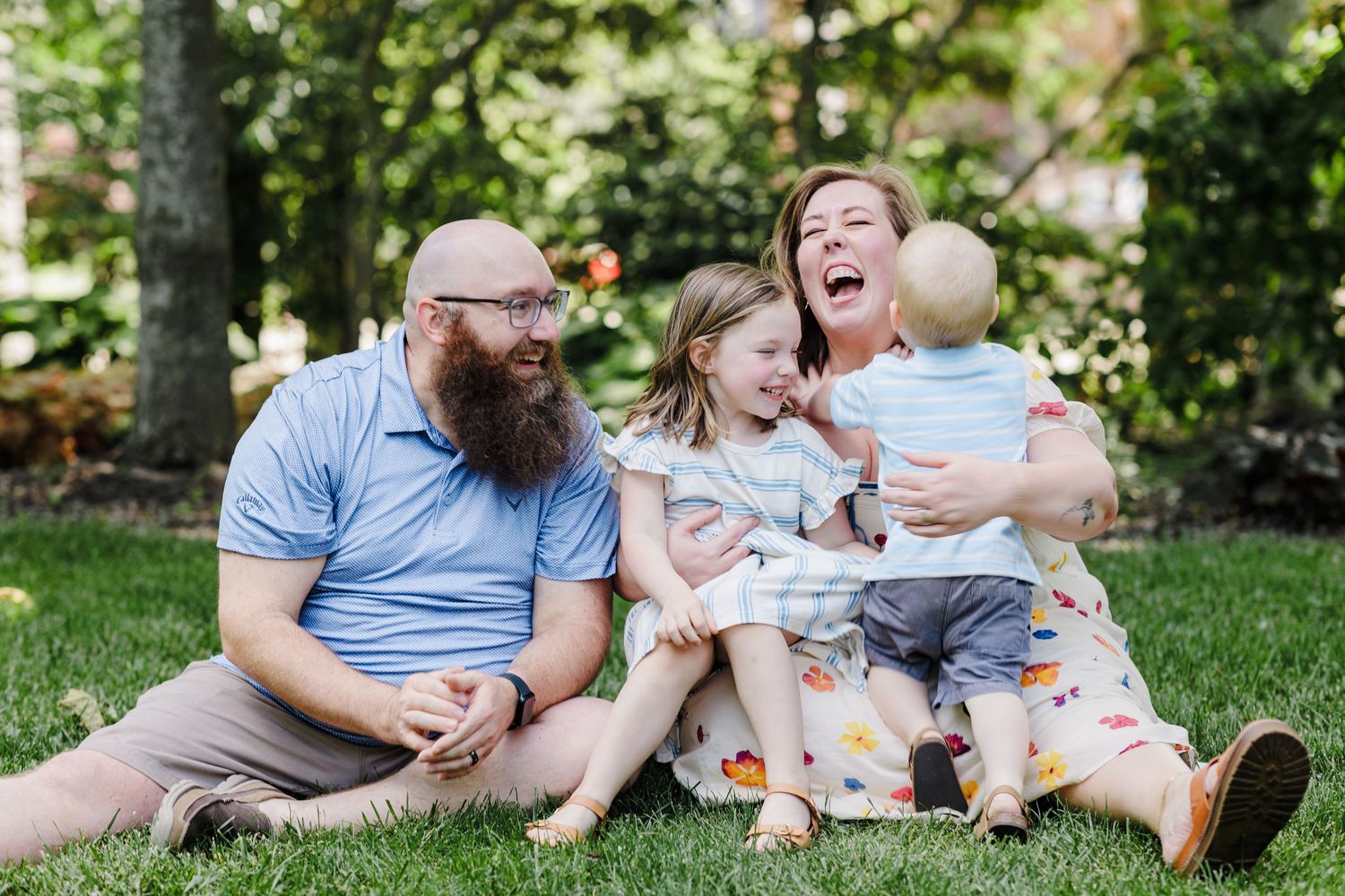 A joyful family of four laughs together while sitting on green grass in a park.