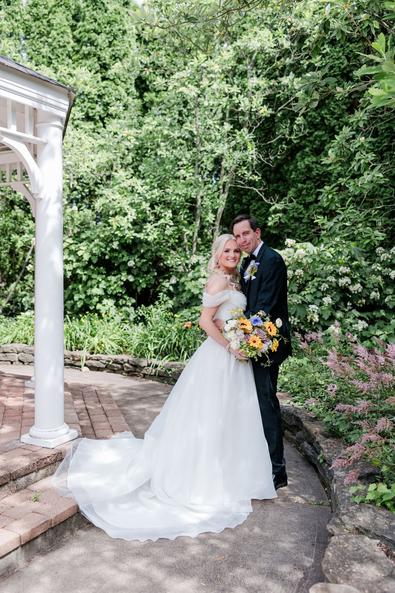 Bride and groom posing near a white gazebo with a lush garden backdrop and sunflower bouquet at the manor house.