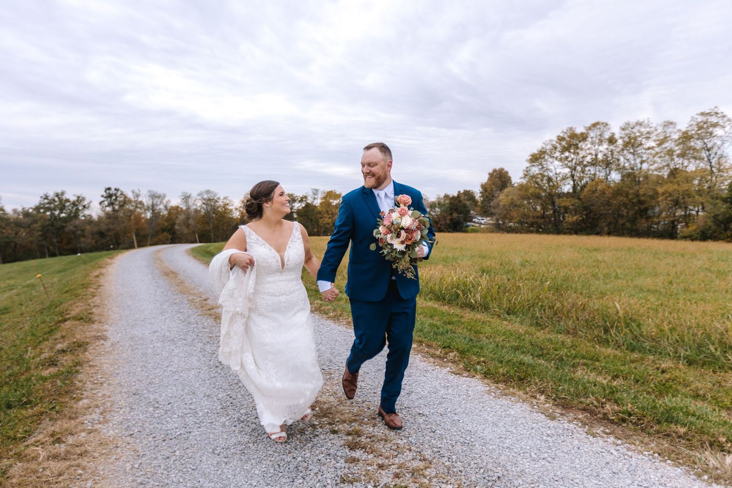 Happy bride and groom walking hand in hand down a gravel path through open countryside fields.