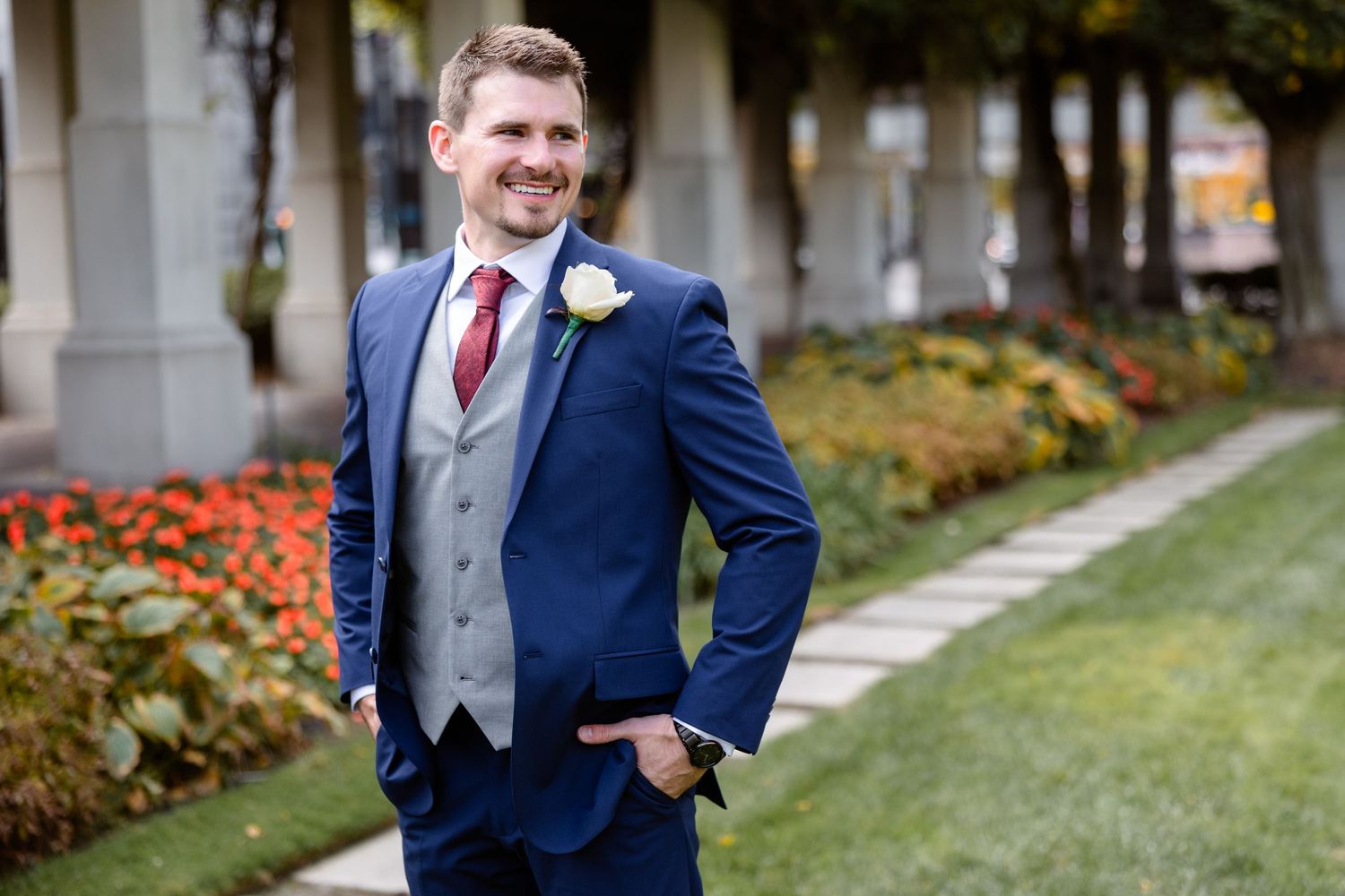 Smiling groom in a navy blue three-piece suit with red tie and white boutonniere posing in a garden.