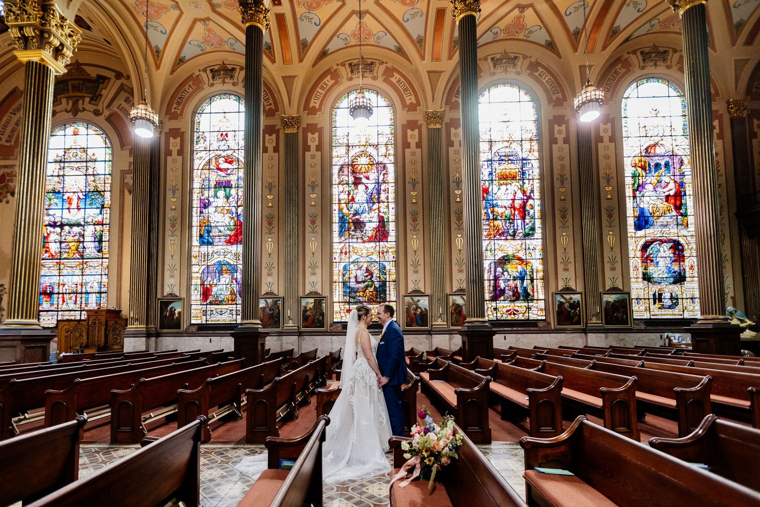 Bride and groom stand in a stunning church aisle surrounded by ornate stained glass windows.