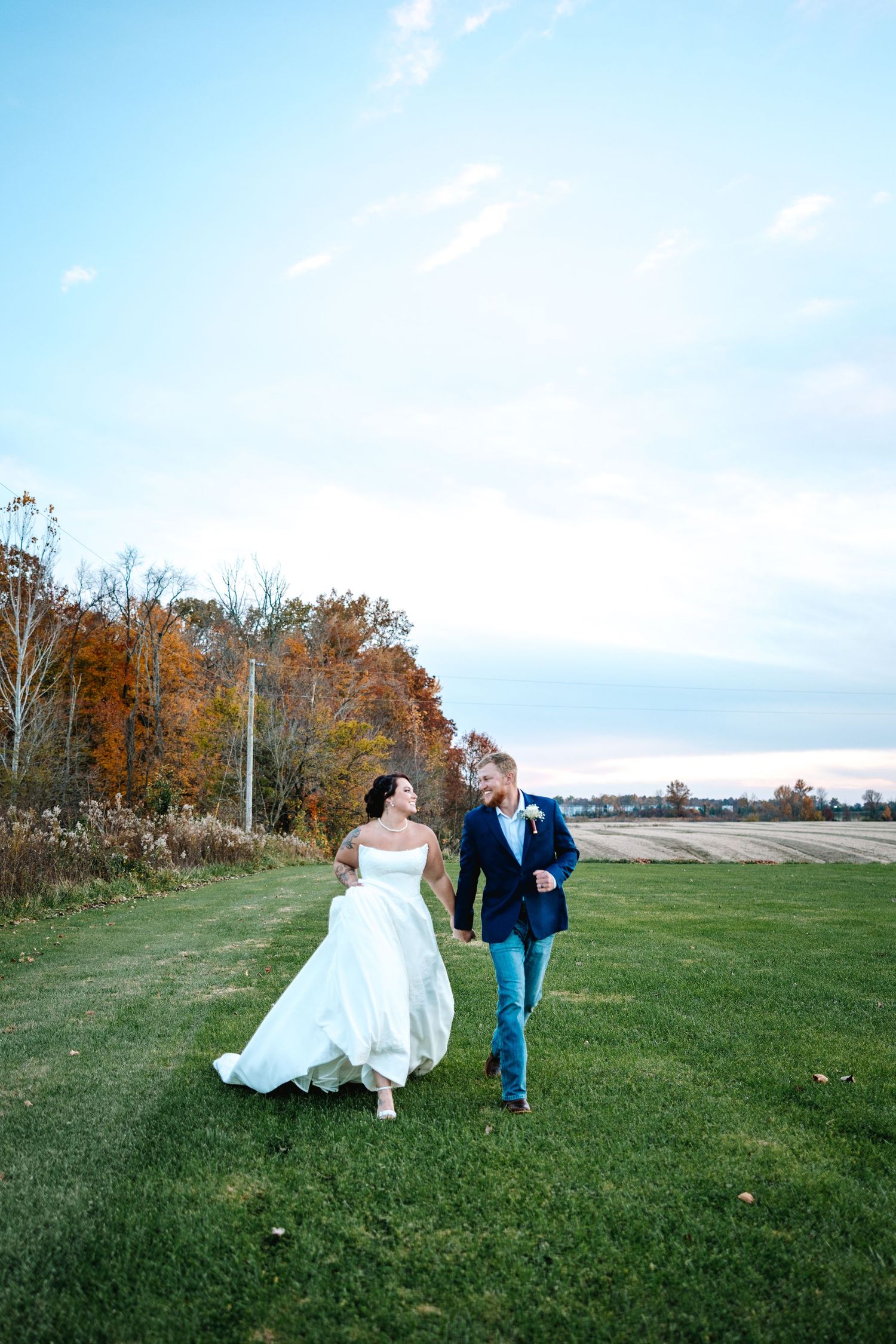 Bride and groom walk hand in hand across a green field with vibrant autumn trees in the background.