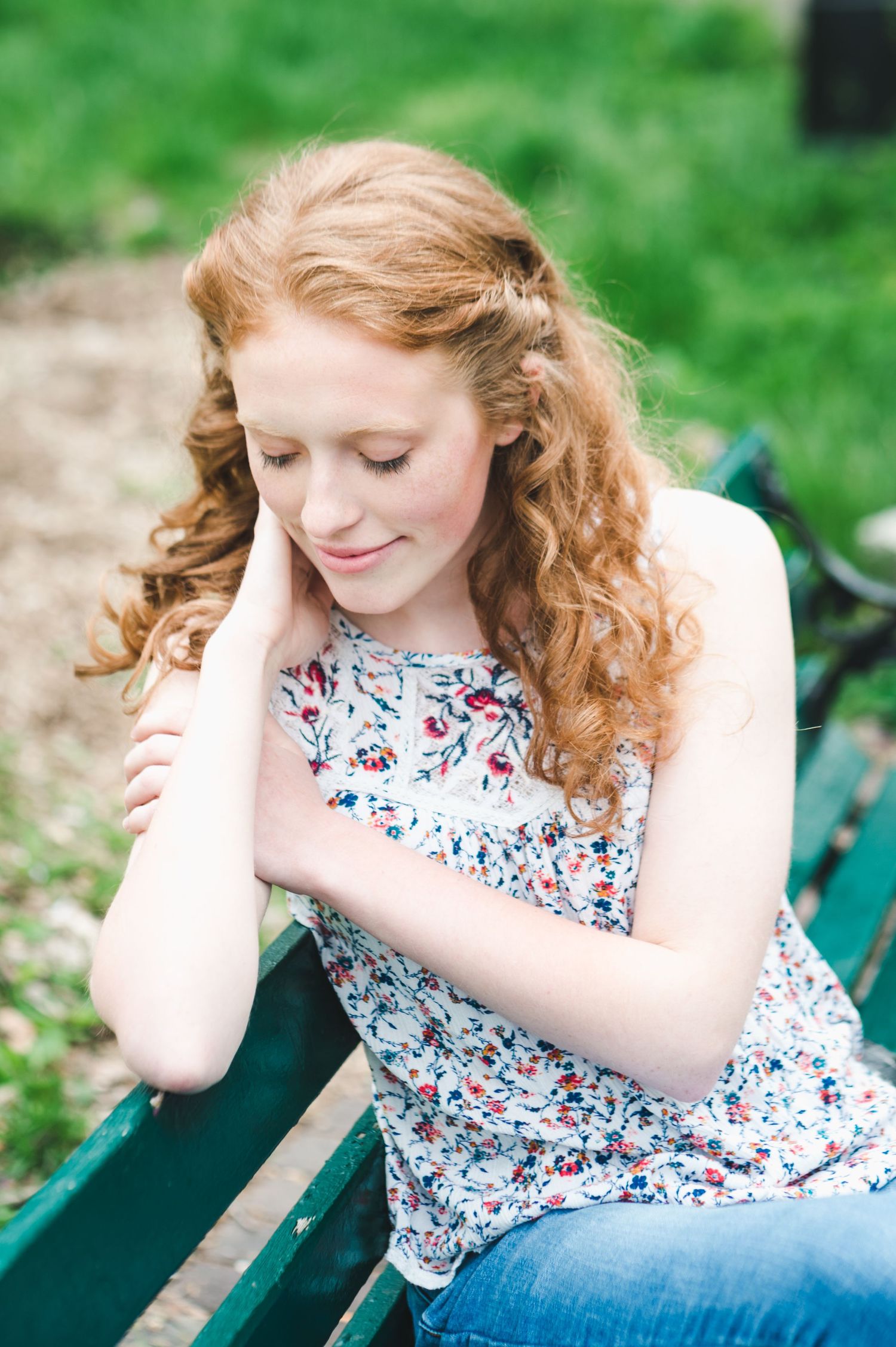 Young woman with curly red hair sits on a green bench, wearing a floral top, looking down softly.