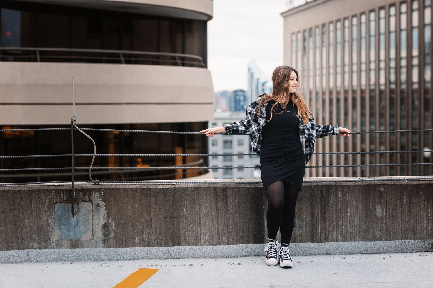 Young woman in black dress and plaid jacket posing on rooftop parking garage with city skyline.