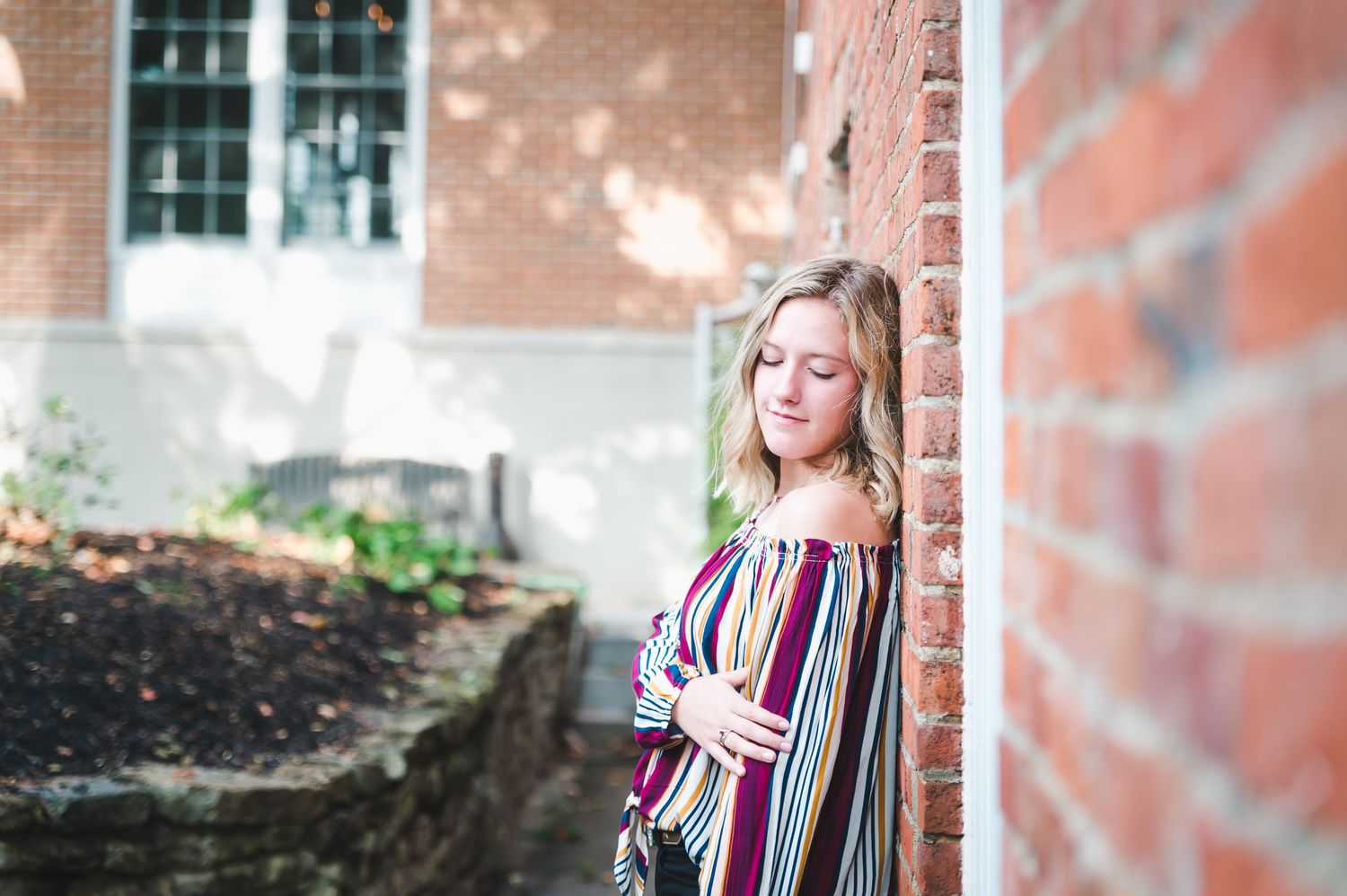 Young blonde woman leans against a red brick wall in a colorful striped off-shoulder top outdoors.