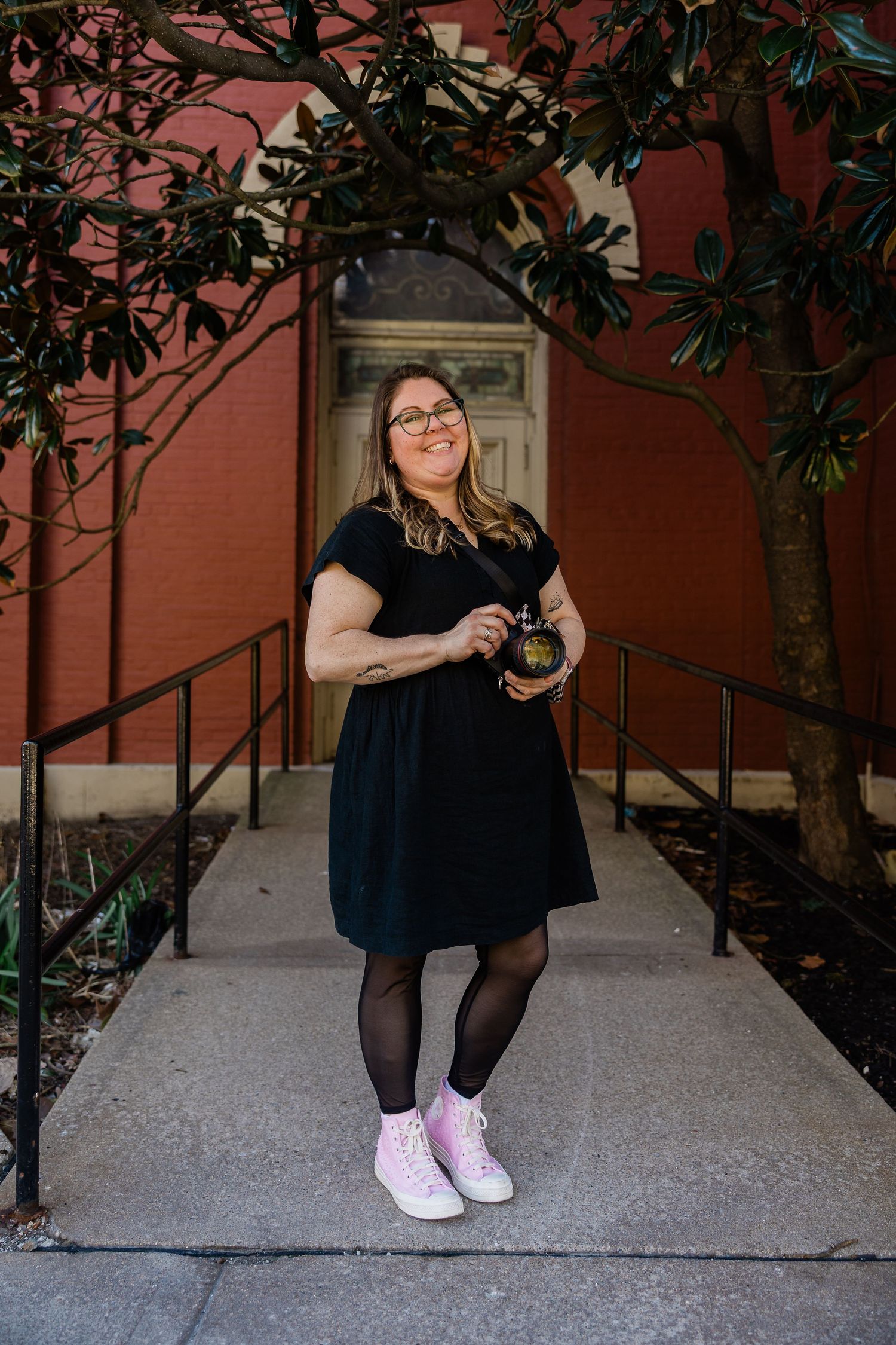 Woman in black dress and pink sneakers smiling while holding camera outside red brick building.