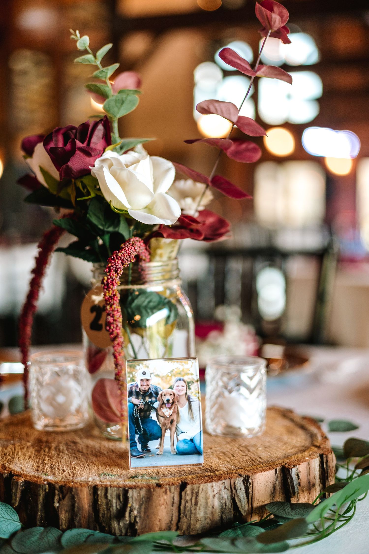 Rustic wedding centerpiece with burgundy flowers in mason jar and couple photo on wood slice.