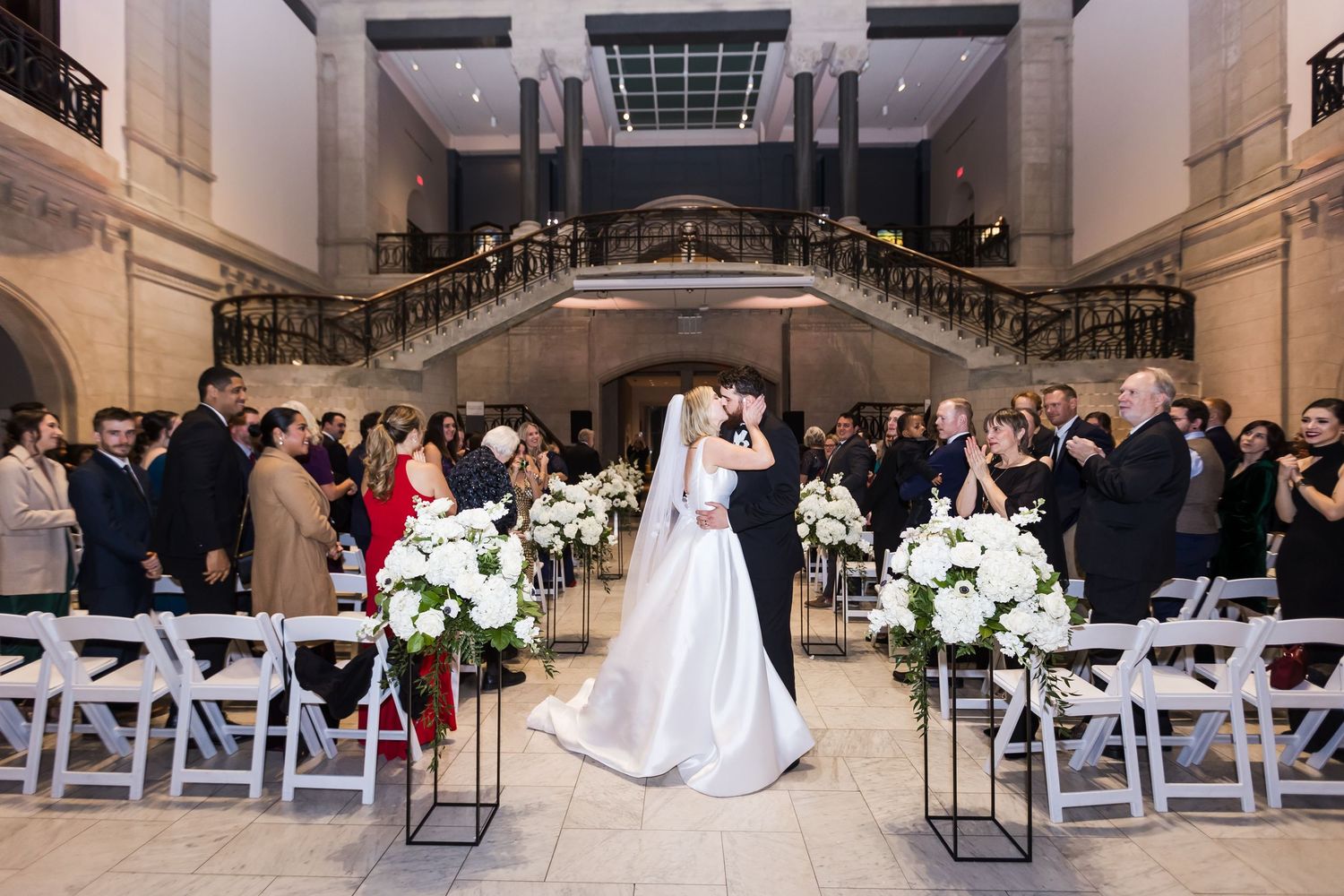 Bride and groom share first kiss at elegant indoor wedding ceremony with white floral arrangements at The Cincinnati Art Museum.