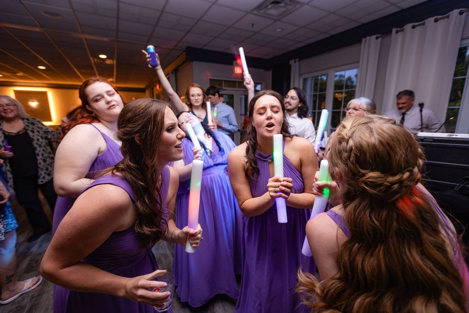 Bridesmaids in purple dresses wave colorful foam sticks while dancing at a wedding reception.