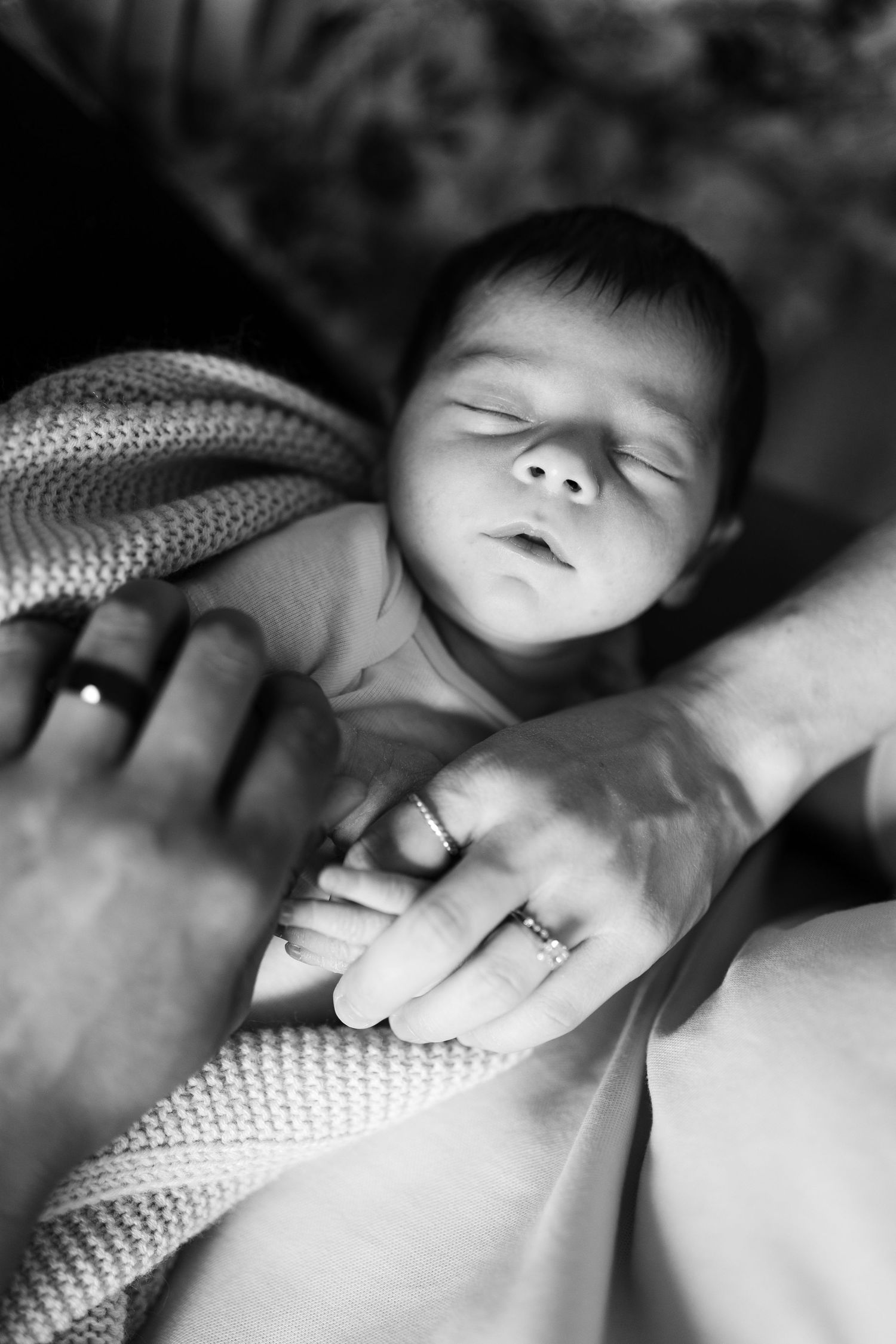 Black and white close-up of a sleeping newborn baby held gently by parents wearing wedding rings.