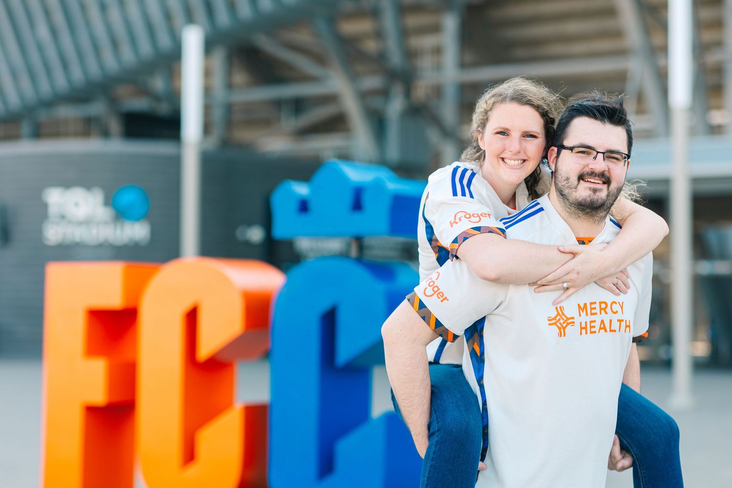 Couple in FC Cincinnati jerseys posing at TQL Stadium colorful sign.