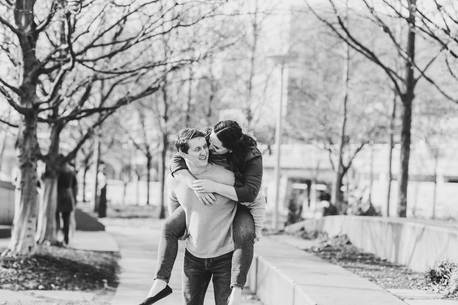 Black and white photo of a couple sharing a piggyback ride on a tree-lined park pathway.