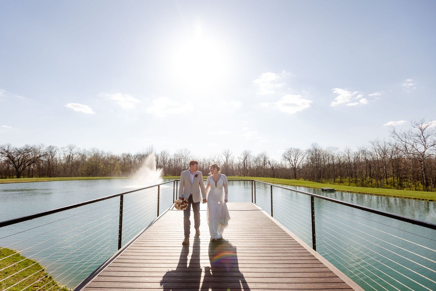 Bride and groom hold hands walking on scenic dock over sparkling lake on sunny wedding day at Hanover Reserve.