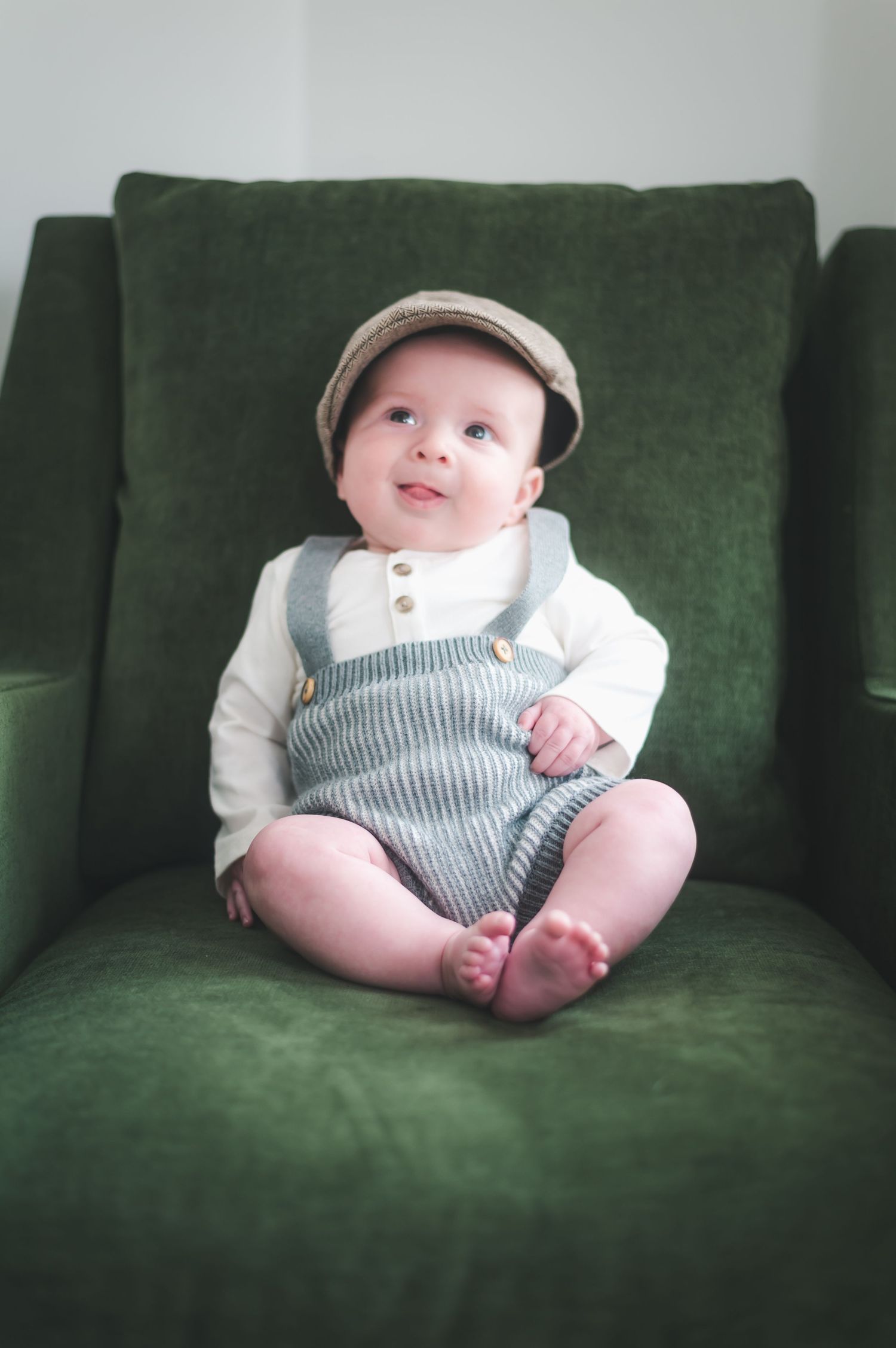 Adorable baby boy in striped overalls and flat cap sitting in a green velvet chair.