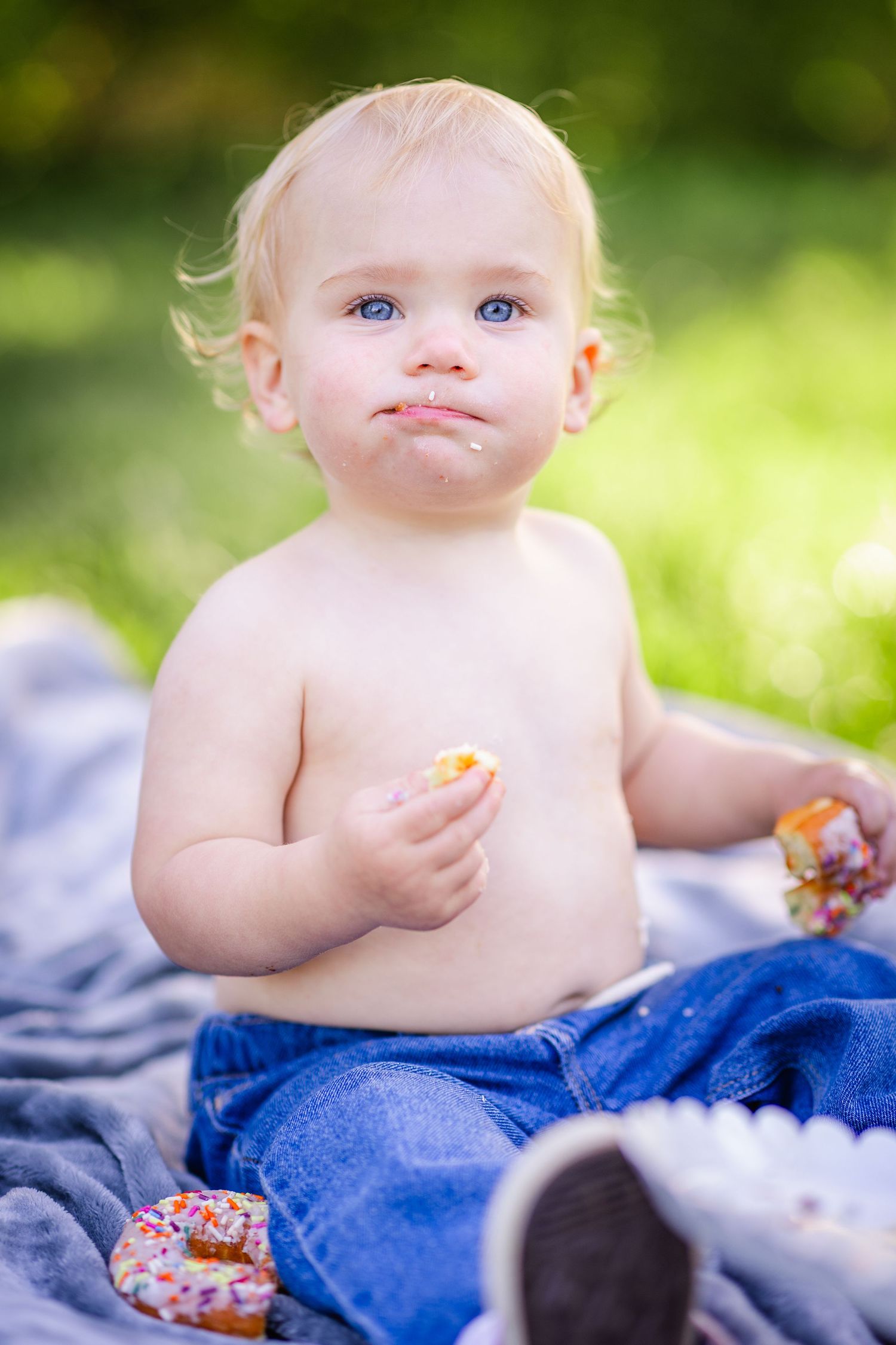 Cute blue-eyed baby with messy face holding sprinkled donuts while sitting outdoors on a blanket.