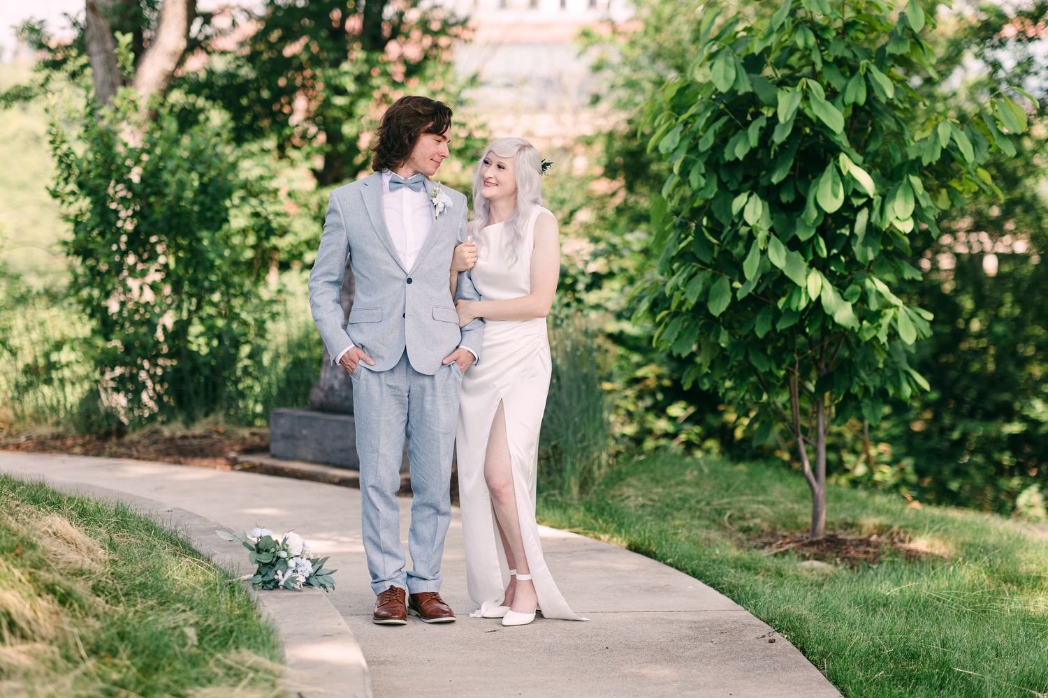 Bride in elegant slit wedding dress and groom in light blue suit share a romantic moment on garden path.
