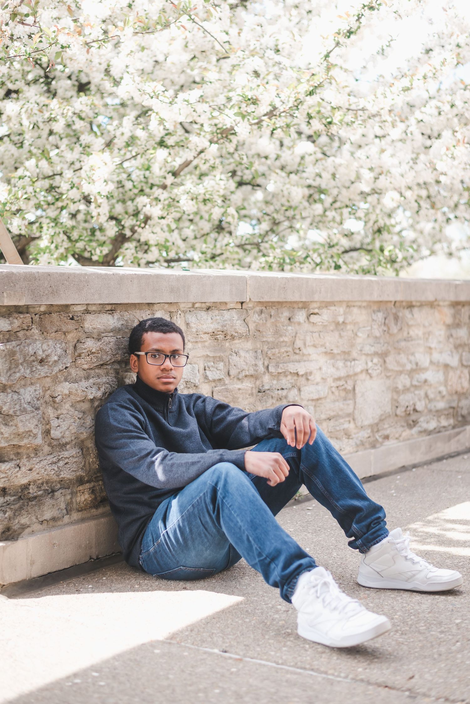 Young man in glasses sitting against stone wall under blooming white flowering tree.