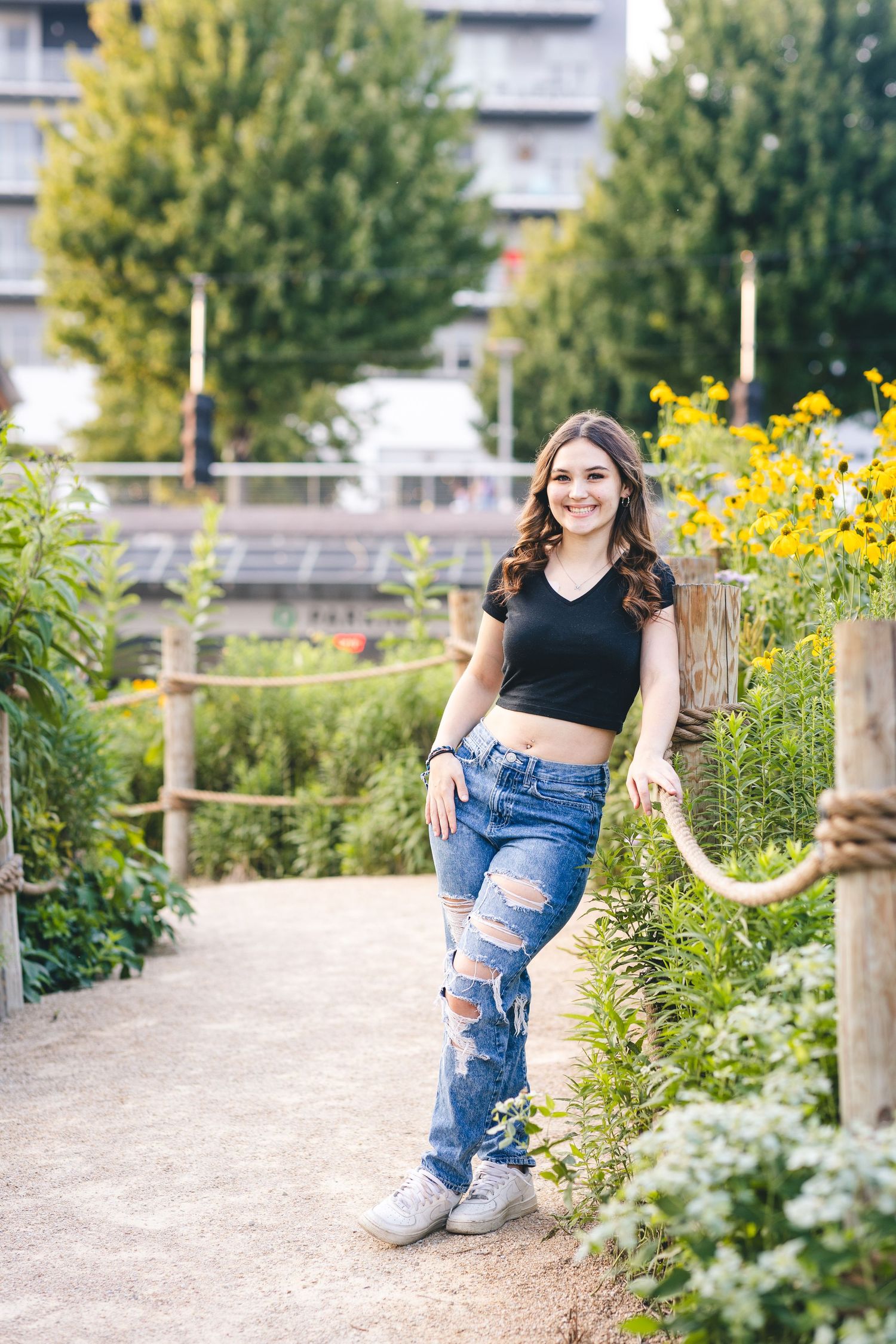 Smiling young woman in black crop top and ripped jeans posing in a lush garden pathway.