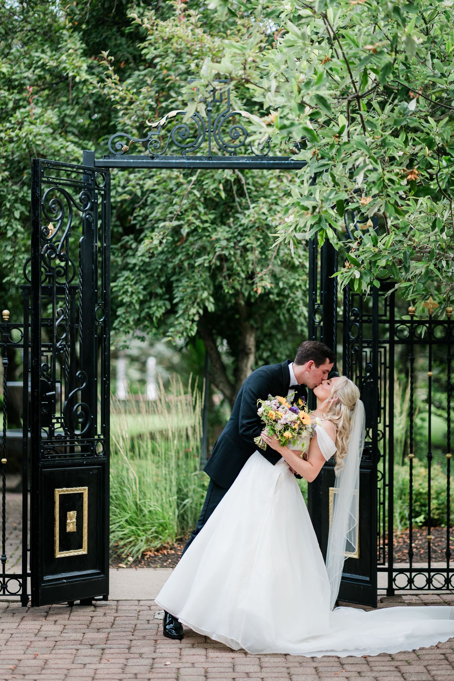 Bride and groom share a romantic dip kiss in front of elegant black iron garden gates surrounded by lush greenery.