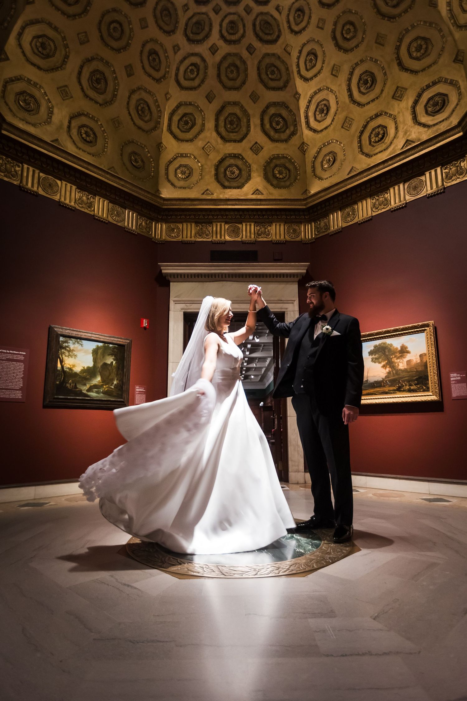 Bride and groom dance beneath ornate gilded ceiling in elegant red-walled Cincinnati art museum gallery.
