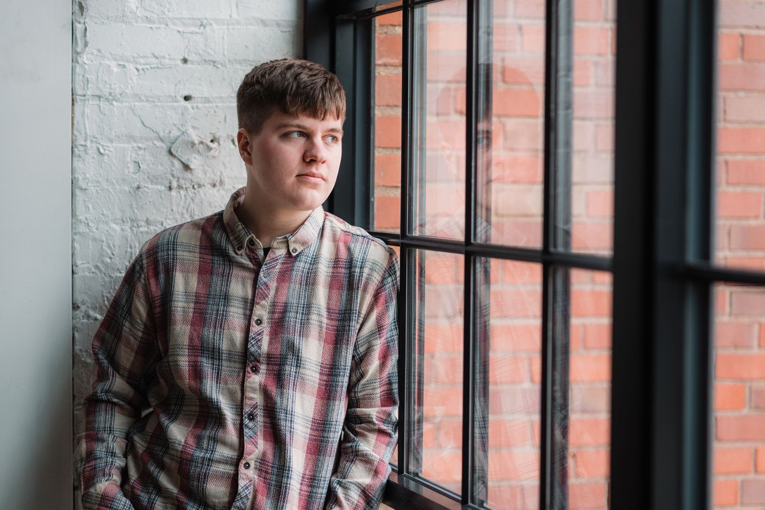 Young man in plaid shirt gazes thoughtfully out large industrial window against brick wall background.