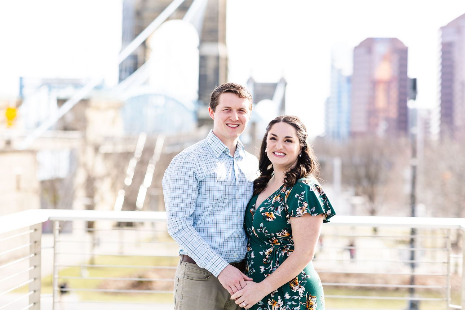 Happy couple posing together on a bridge with Roebling Bridge and skyline in the background.