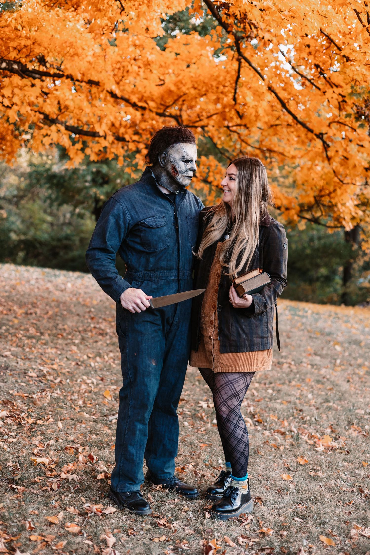 Couple poses in autumn park, man in Michael Myers Halloween costume with mask and knife beside smiling woman.