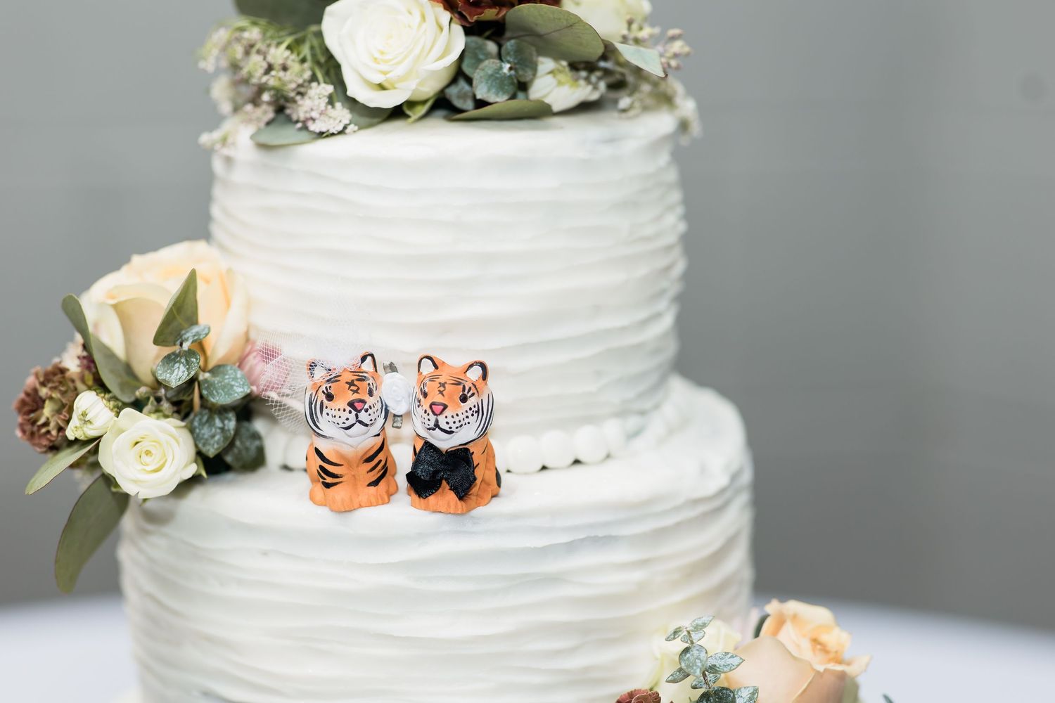 Three-tier white textured wedding cake topped with cute tiger figurines and floral decorations.