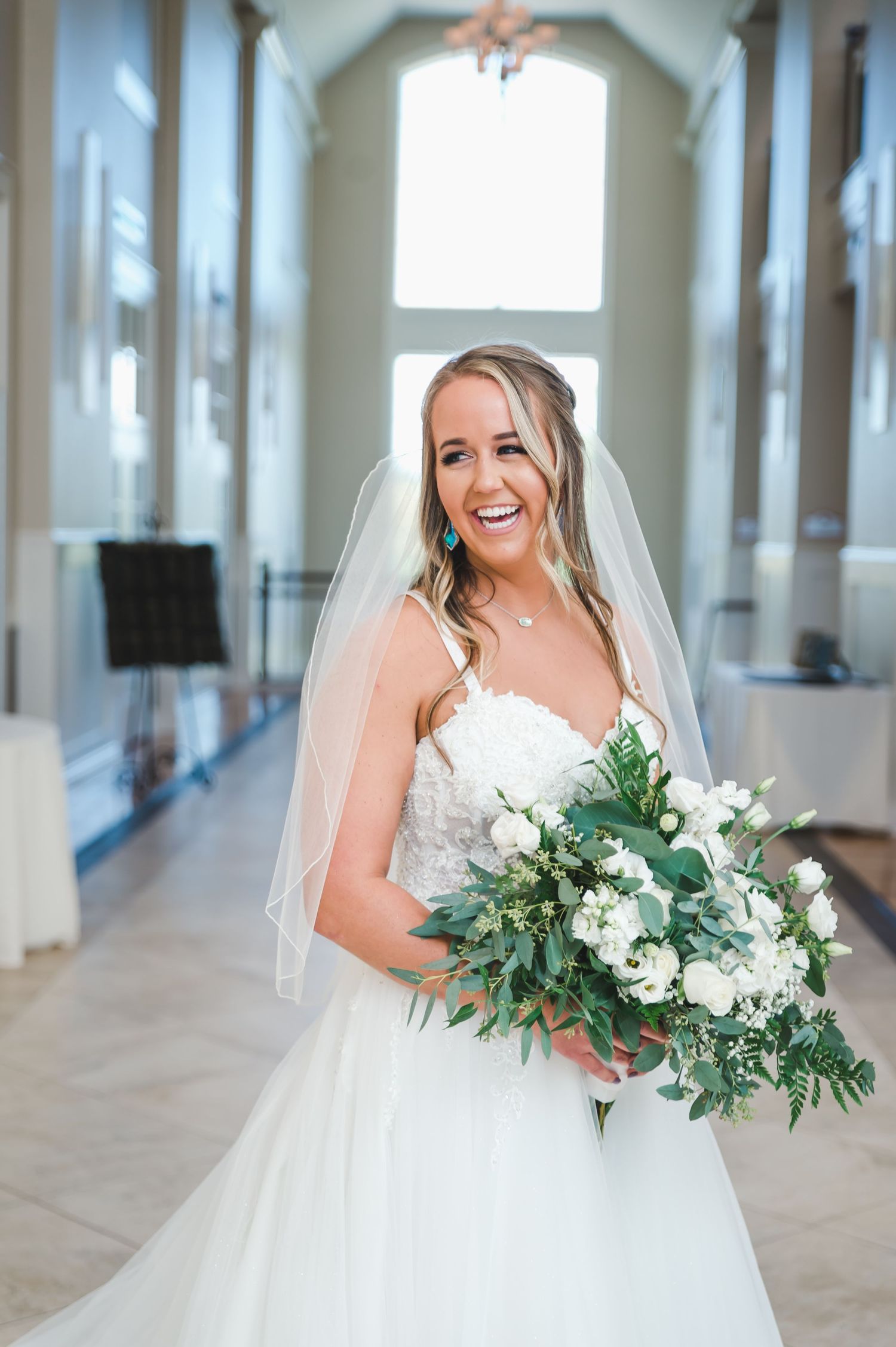 Smiling blonde bride in lace gown with veil holding large white and greenery bouquet indoors.