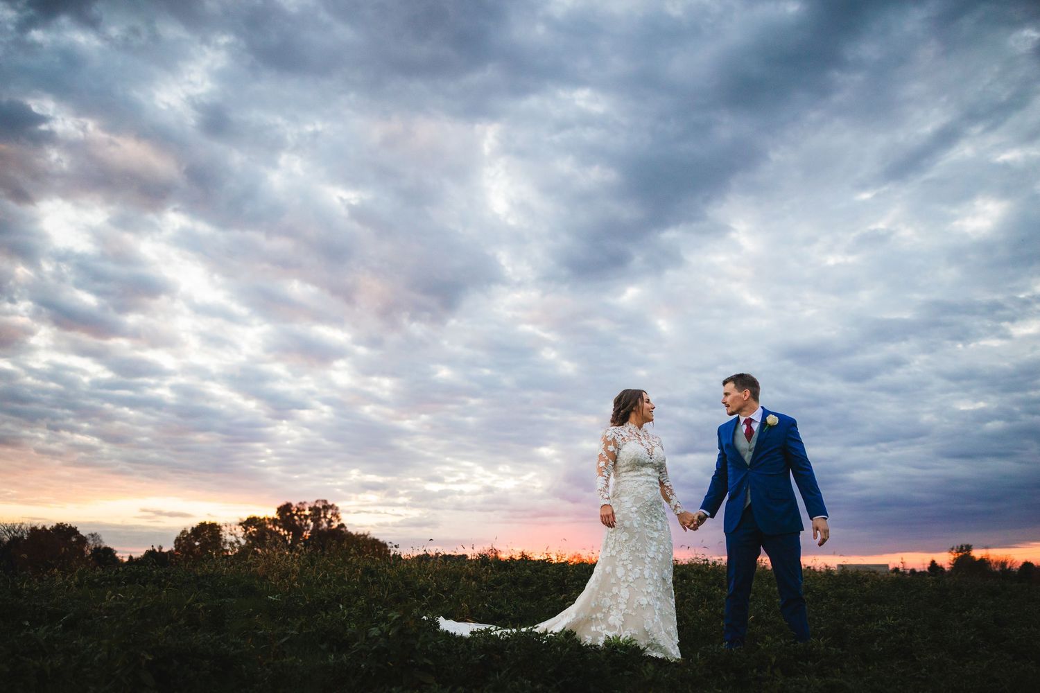 Bride and groom hold hands at sunset under dramatic cloudy sky in an outdoor wedding portrait.
