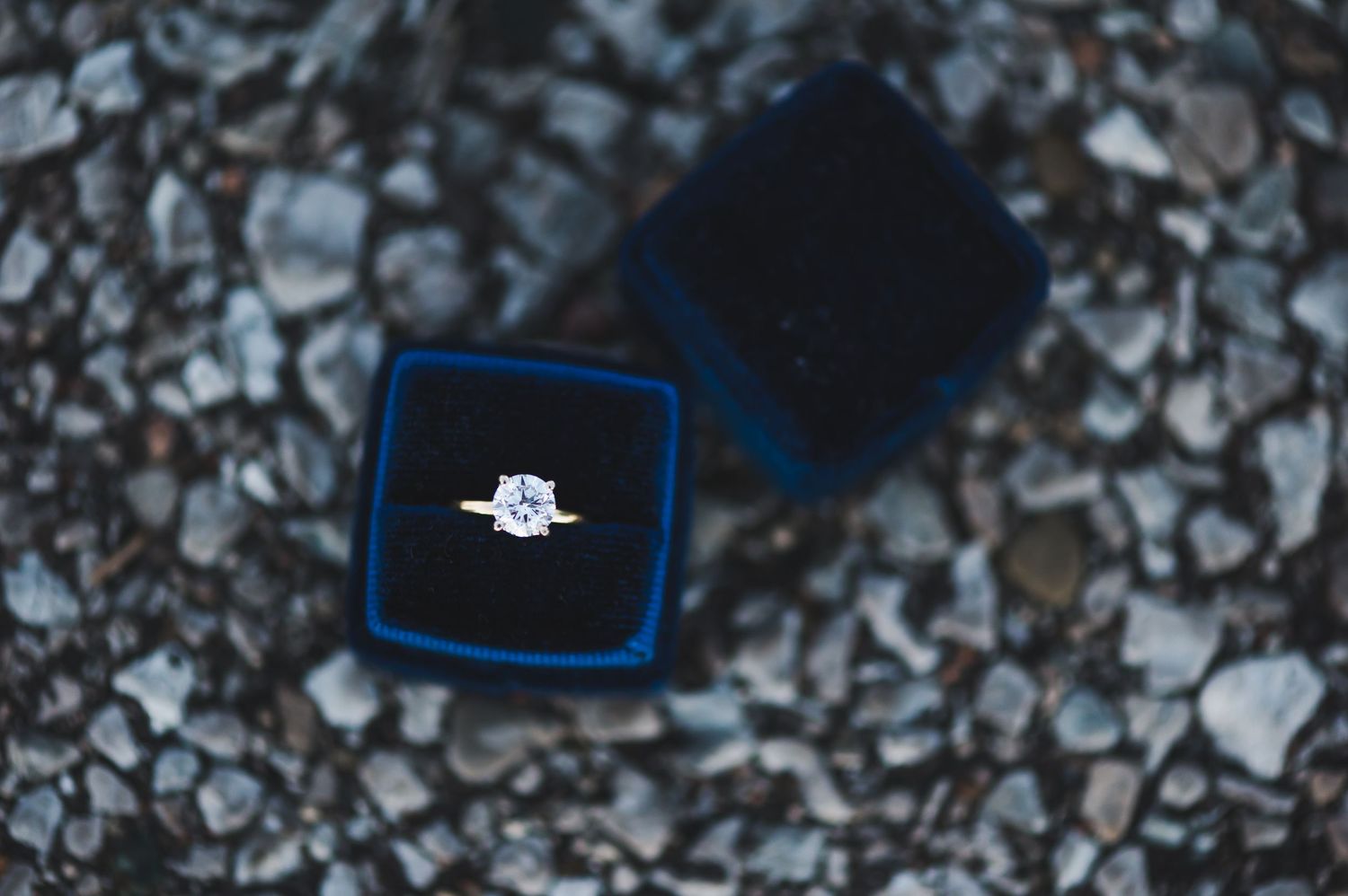 Diamond solitaire engagement ring in blue velvet box displayed on rocky stone surface.